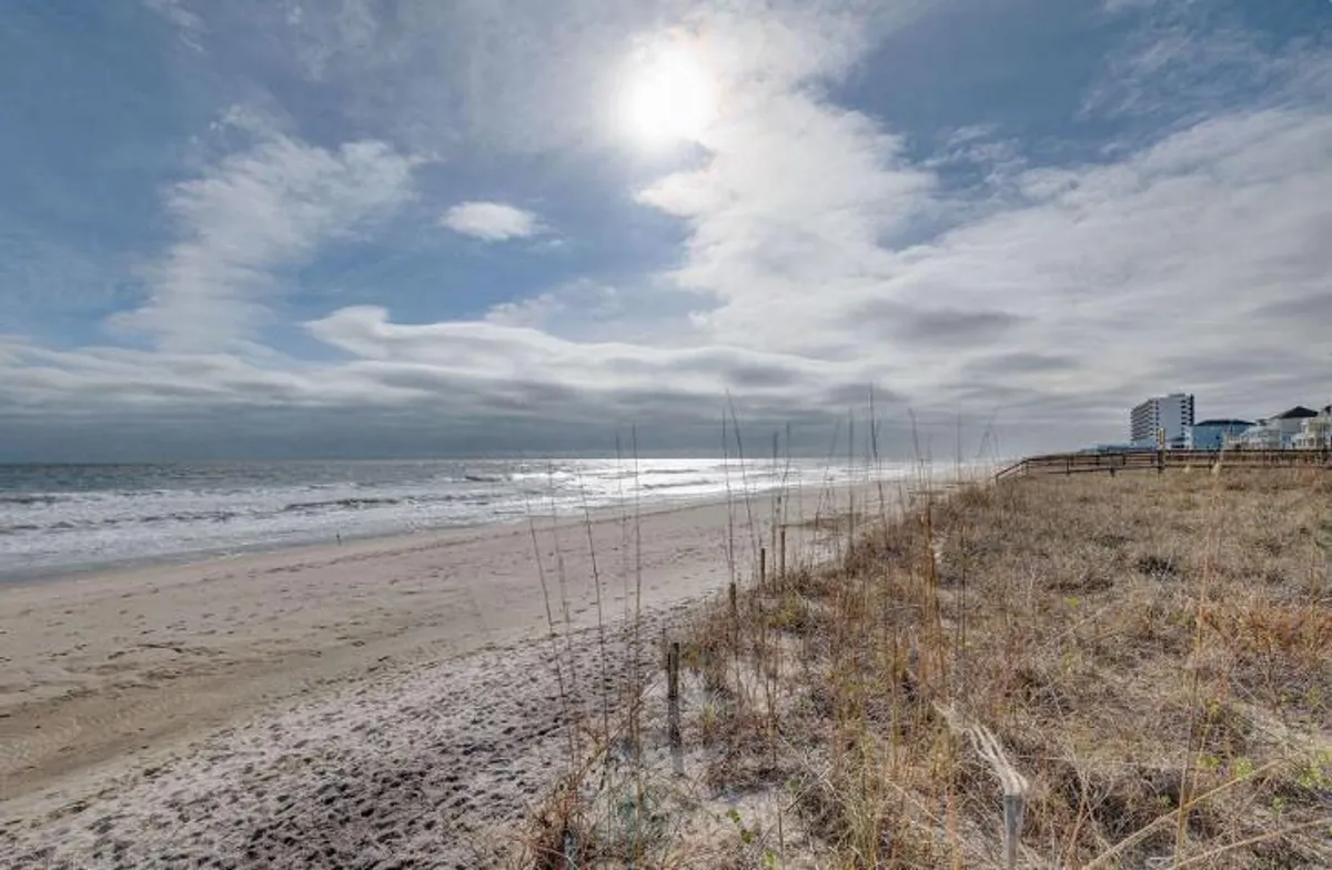 A wide shot of the sandy shoreline, ocean waves, dune grass, and sunlight streaming through the clouds. The beach is inviting and active, perfect for surfing, sunbathing, or simply soaking up the coastal vibe with friends.