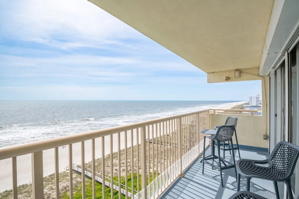 A balcony scene overlooking the sweeping shoreline, with comfortable seating positioned to take in expansive ocean views. There's a tranquil mood, with the ocean stretching into the distance and hints of guests relaxing, perhaps with a book or morning coffee.