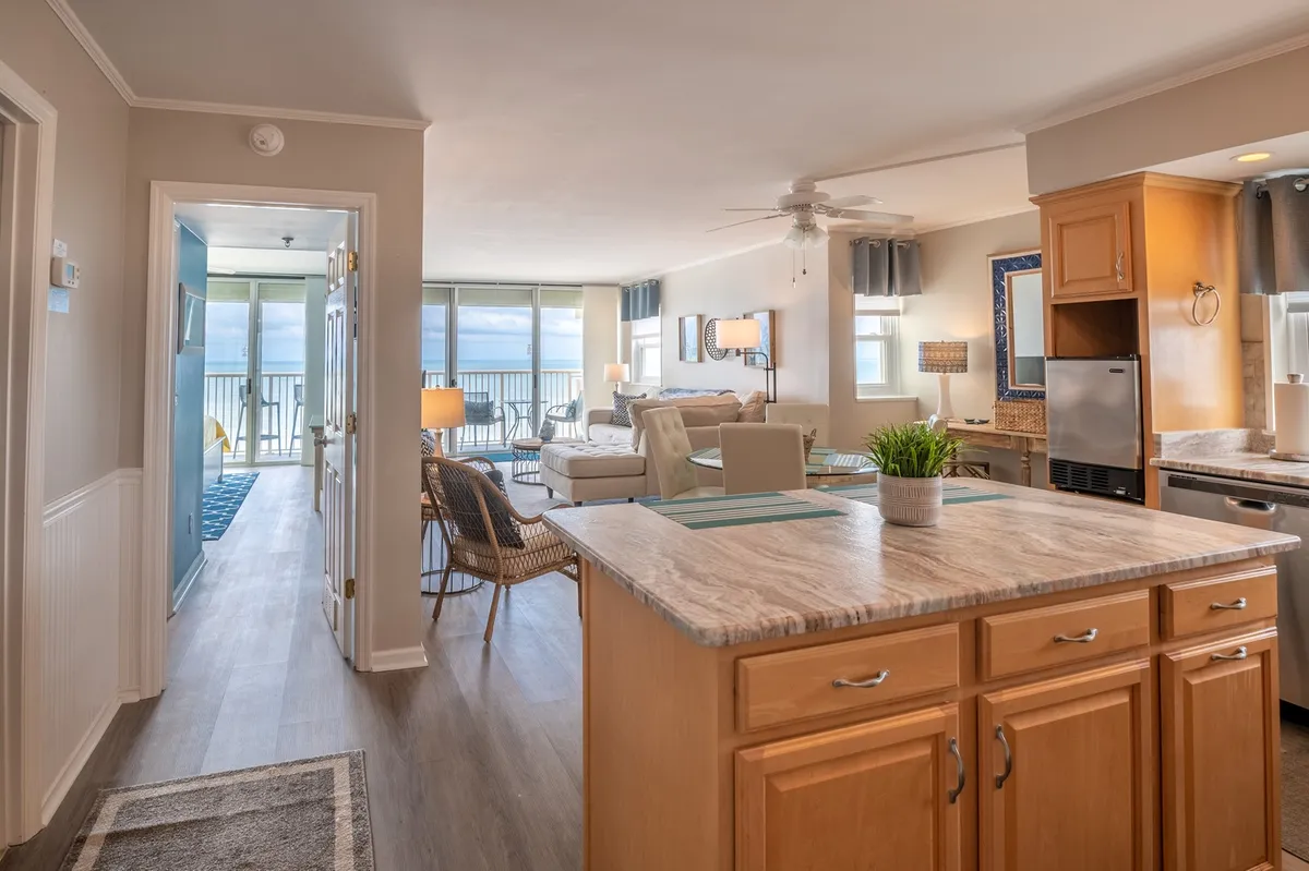 An open-concept kitchen with a central island that transitions seamlessly into a living area, with a balcony view in the background. The scene suggests a space filled with natural light, perfect for both meal prep and socializing, with views that connect kitchen activities to the broader living experience.