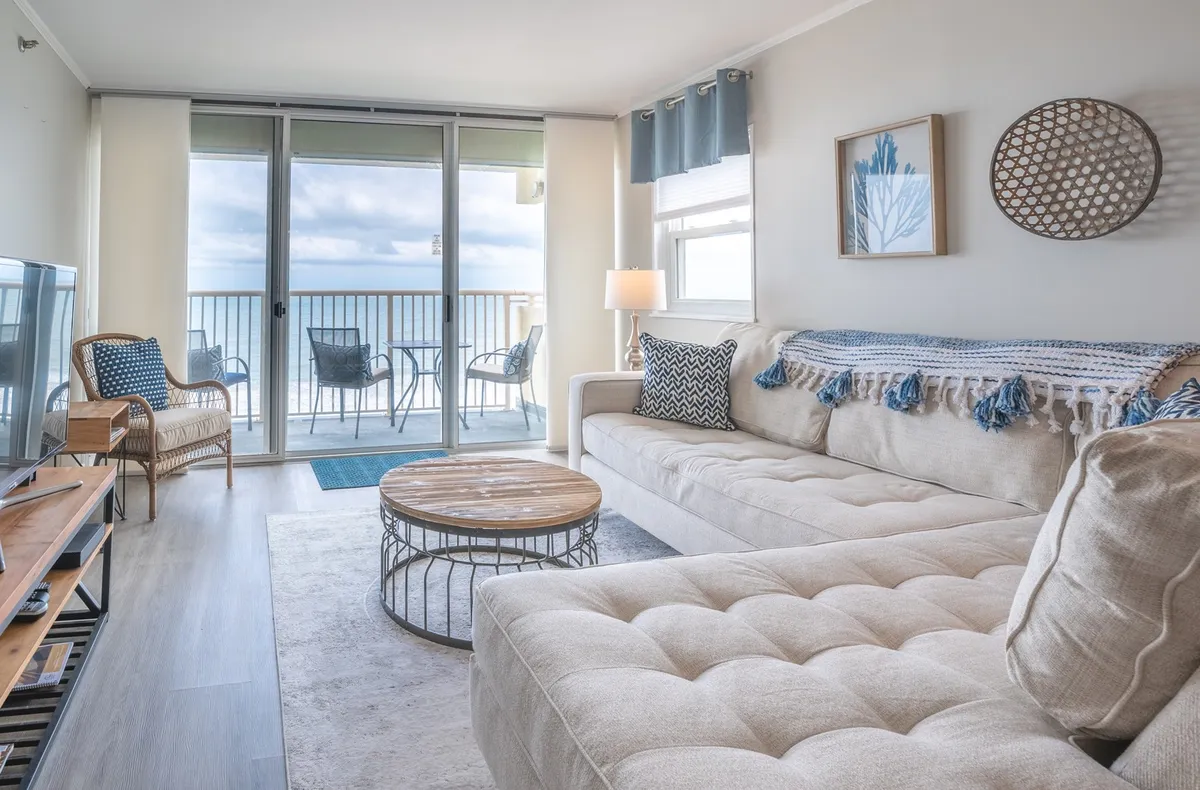 The image showcases a bright, airy living room featuring a large, comfortable sectional sofa and a coffee table, all arranged to face a sliding glass door that opens to a stunning ocean view. Natural light floods the space, inviting guests to relax indoors while still enjoying the beauty of the coastline.