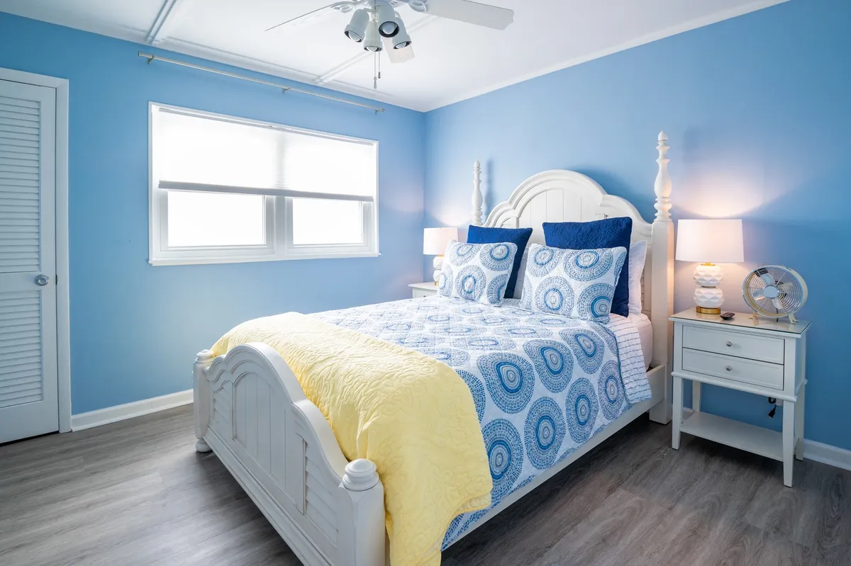 A welcoming bedroom with a queen-sized bed adorned in blue and yellow bedding, flanked by white nightstands. A large window fills the room with daylight, creating a refreshing and restful environment.