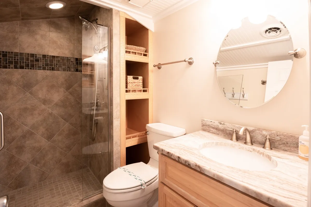 A modern bathroom with a glass shower enclosure, marble countertop, and wooden shelves stocked with fresh linens. The space feels clean, serene, and ideal for self-care routines or a refreshing shower after a day at the beach.