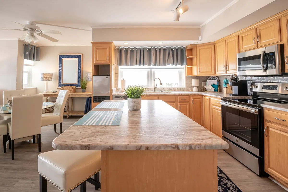 A welcoming kitchen scene with a prominent island at the center, surrounded by stainless steel appliances and large windows with curtains. The setup is ideal for cooking and entertaining, offering space for meal prep and gatherings. The overall mood is cozy and inviting, reinforcing the kitchen as a social hub.