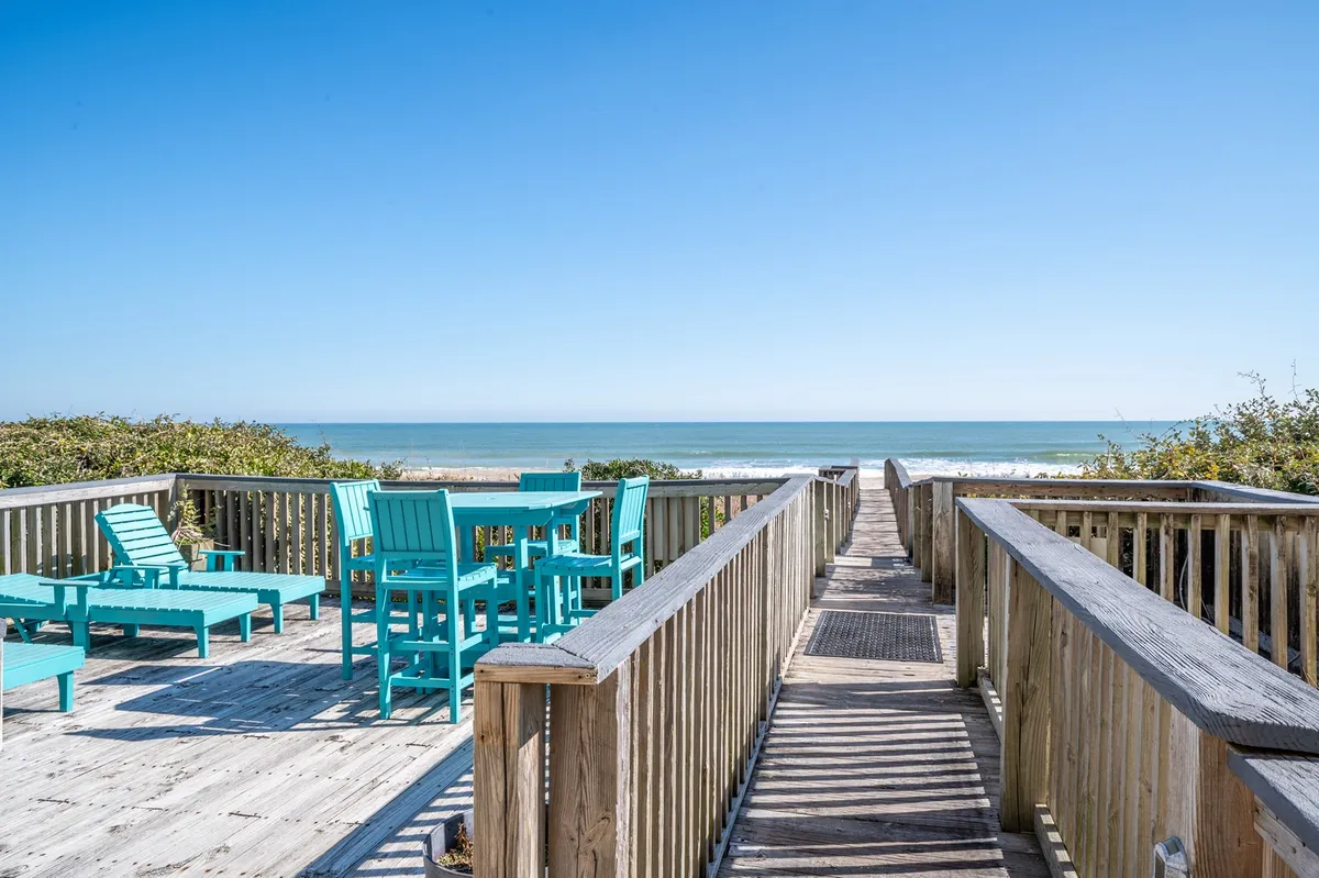 This exterior image features turquoise outdoor furniture on a patio, a boardwalk leading directly to the sandy beach, and a panoramic ocean view. The inviting setup suggests easy transitions between lounging, dining, and playing outdoors, with the boardwalk emphasizing convenient beach access for families.