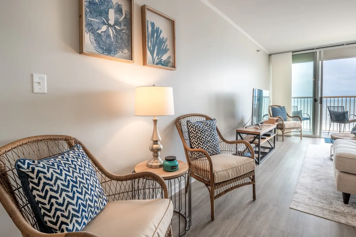 A cozy seating area with wicker chairs, a stylish lamp, artwork on the wall, and a TV, all oriented to enjoy both the calming interior and a view onto the balcony. The balcony doors let in natural light and offer glimpses of the ocean beyond, while the TV and inviting seating set the stage for movie nights or morning relaxation.