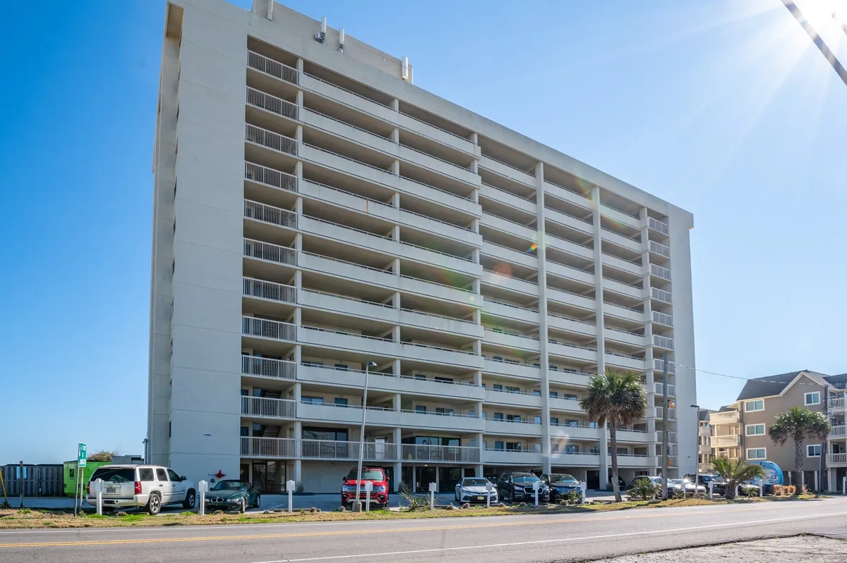 This exterior image showcases the tall Pelican Watch building with multiple balconies, surrounded by palm trees and vehicles, conveying a lively, beachside atmosphere. The visible balconies hint at private outdoor spaces ideal for socializing or unwinding after a day of adventure.