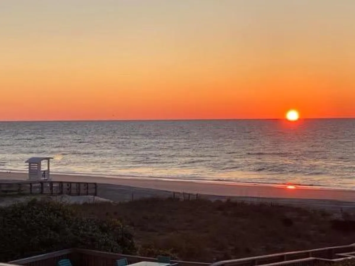 A beautiful view of the sun setting over the ocean, with a distant lifeguard stand and broad sandy beach, creating a tranquil, romantic atmosphere perfect for evening walks or quiet moments together.