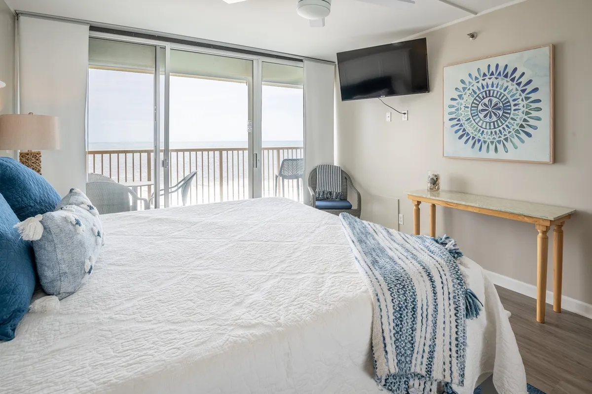 A serene primary bedroom featuring a king-sized bed dressed in crisp white bedding with blue accent pillows. Large windows let in natural light and provide an unobstructed ocean view, while a TV is mounted on the wall for entertainment. A decorative art piece adds a personal touch, and the room opens onto a balcony shared with the living room.