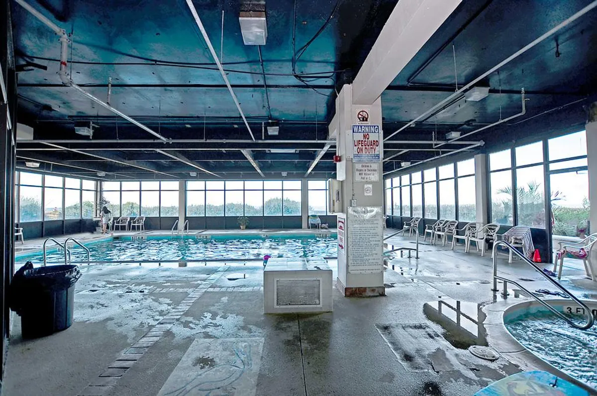 An inviting indoor pool area featuring a large swimming pool, adjacent hot tub, and abundant natural light streaming through expansive windows. Lounge chairs line the poolside, offering a serene spot to relax or socialize after a swim or soak.